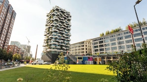 Un paisaje urbano moderno con un edificio de varios niveles distintivo, con balcones, rodeado de otros edificios altos y un área verde bien cuidada.
