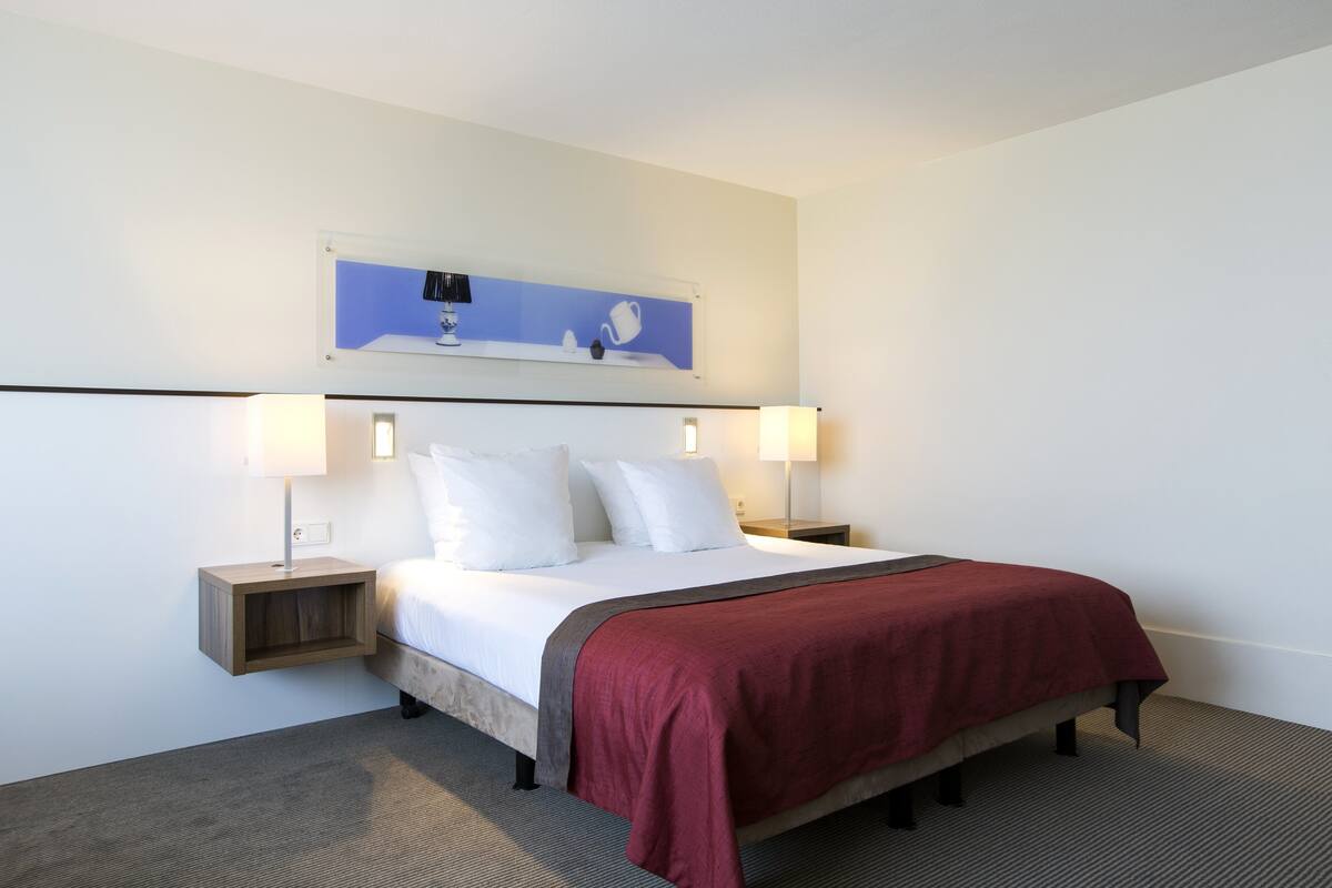 A hotel room with a bed, two bedside tables, a nightstand, a red throw blanket, and a wall-mounted artwork.