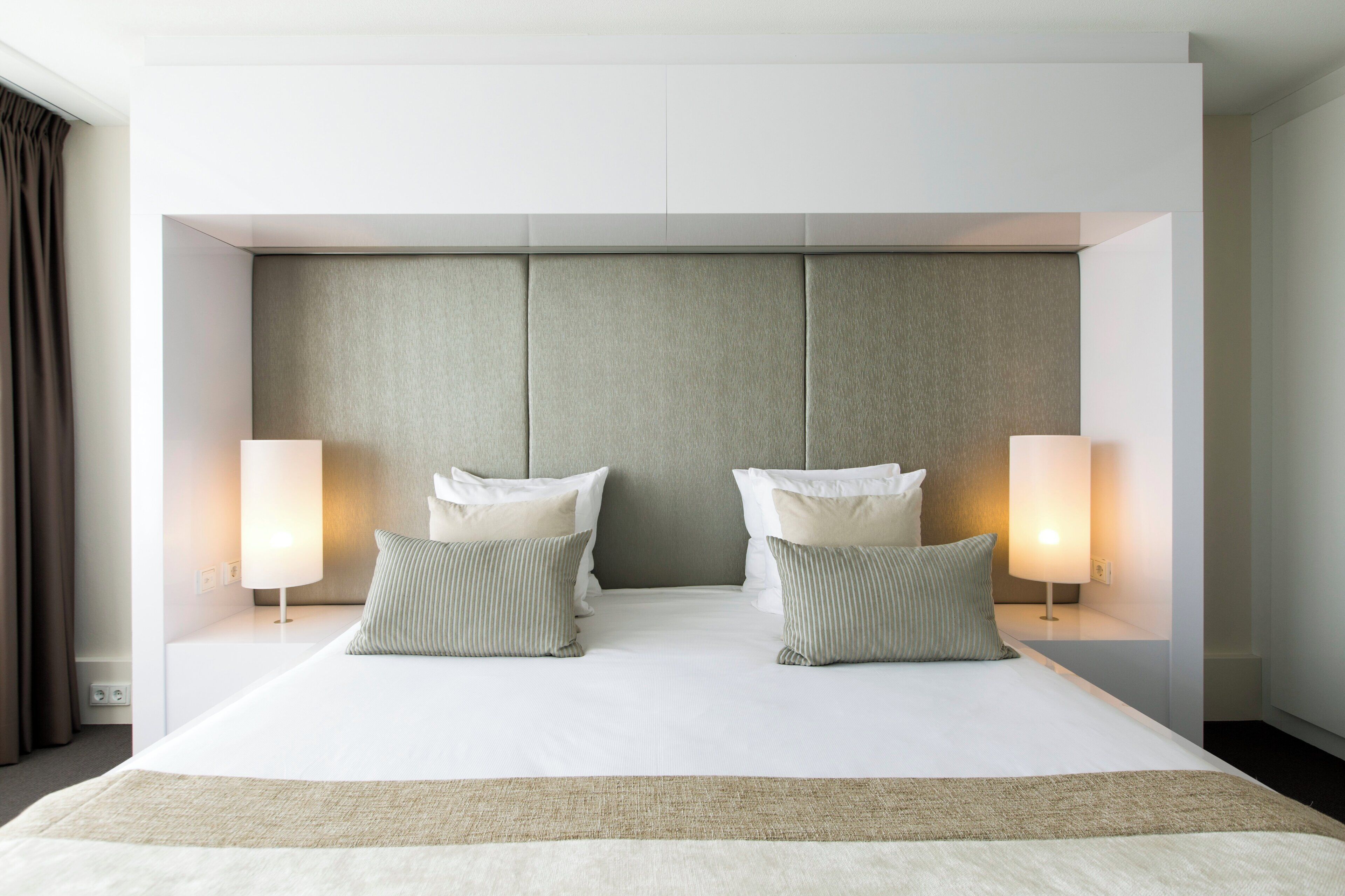 A modern hotel room with a large bed, two bedside lamps, and a neutral-toned headboard.