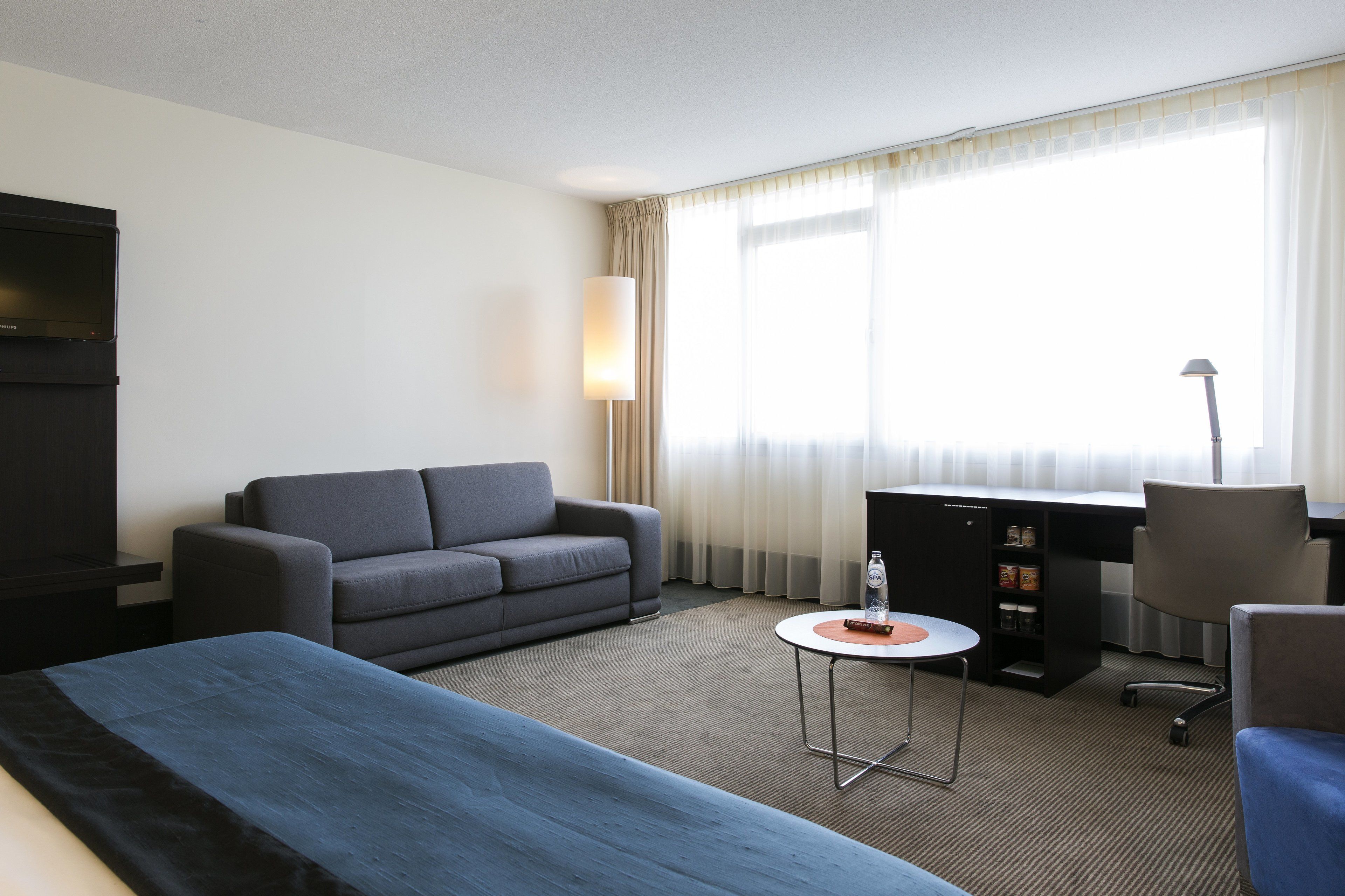 A hotel room with a bed, a sofa, a desk with a computer, a lamp, and a window with curtains.