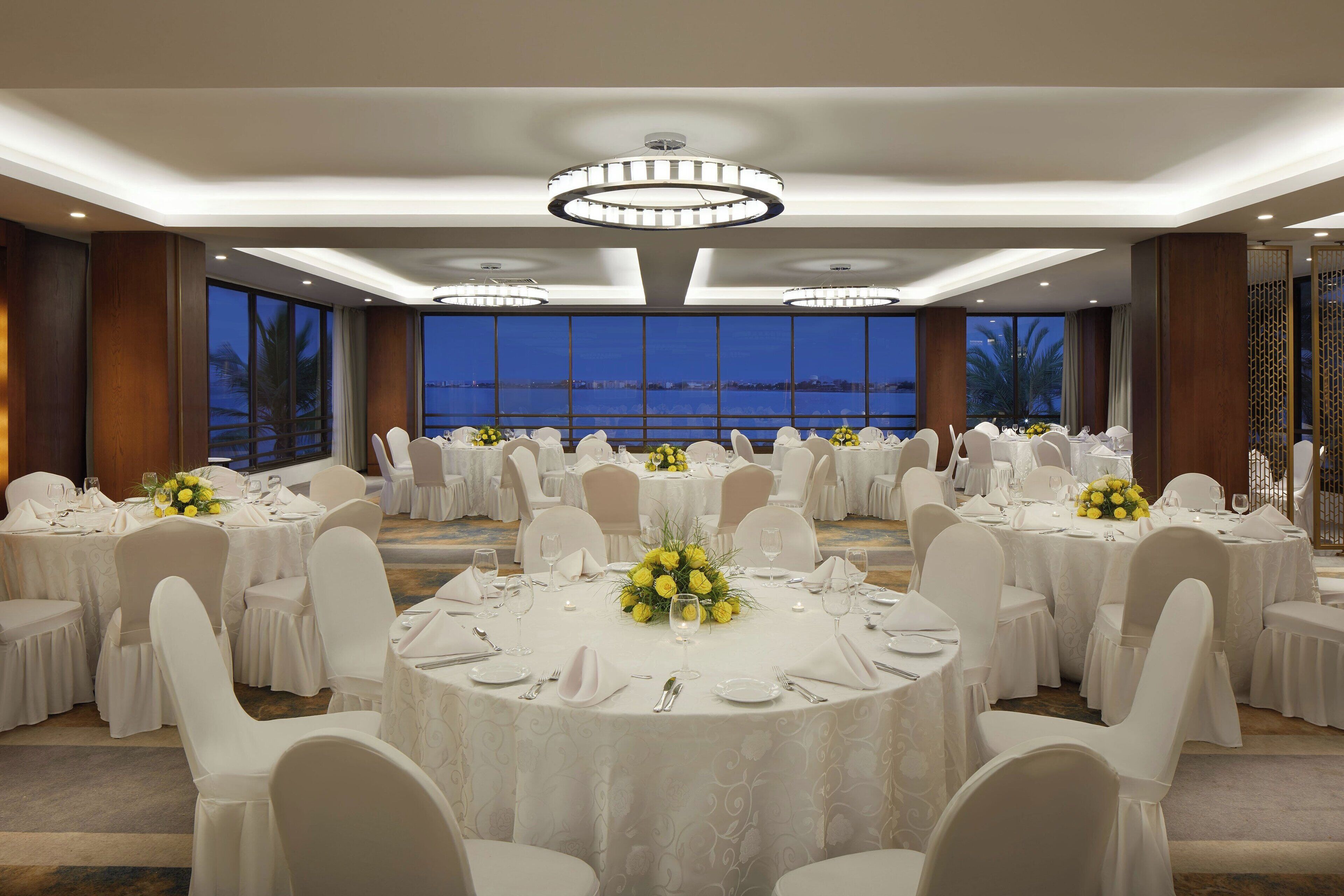 Un salón de banquetes con mesas redondas, sillas blancas y centros de mesa florales amarillos.