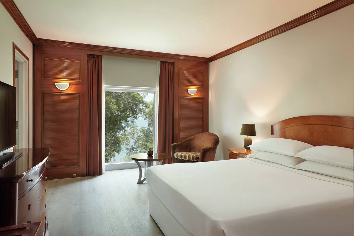 Una habitación de hotel con una cama grande, televisión, silla, mesita y lámpara.