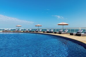 Ein Swimmingpool mit Liegestühlen und Sonnenschirmen, ein klarer Himmel und Blick auf das Meer.