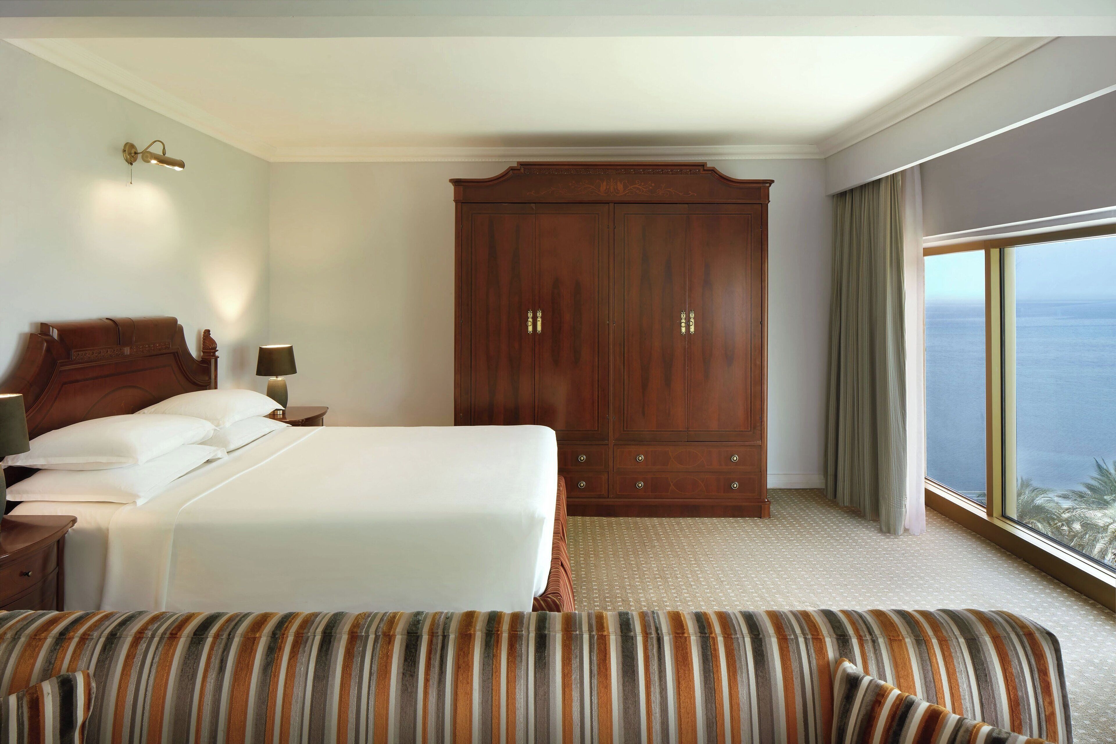 Ein Hotelzimmer mit einem großen Bett, einem Holzschrank, einem Nachttisch mit Lampe und Blick auf das Meer durch ein großes Fenster.