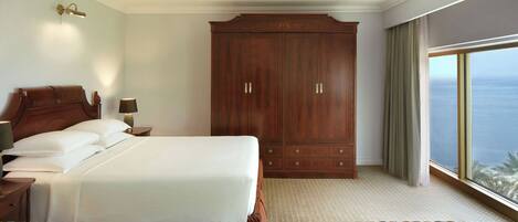 Ein Hotelzimmer mit einem großen Bett, einem Holzschrank, einem Nachttisch mit Lampe und Blick auf das Meer durch ein großes Fenster.
