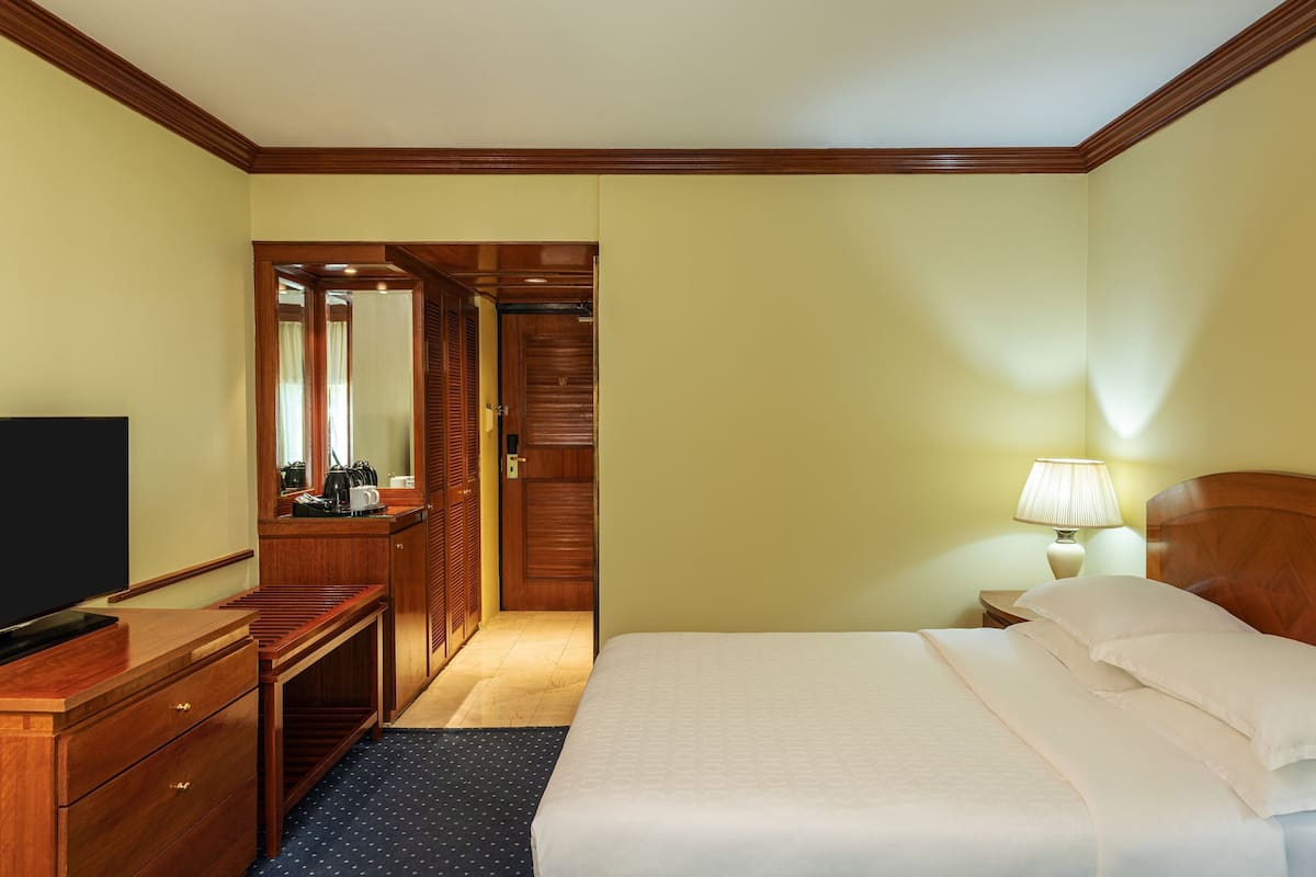Una habitación de hotel con una cama, un escritorio con televisor, un espejo, un armario y una mesita de noche con lámpara.