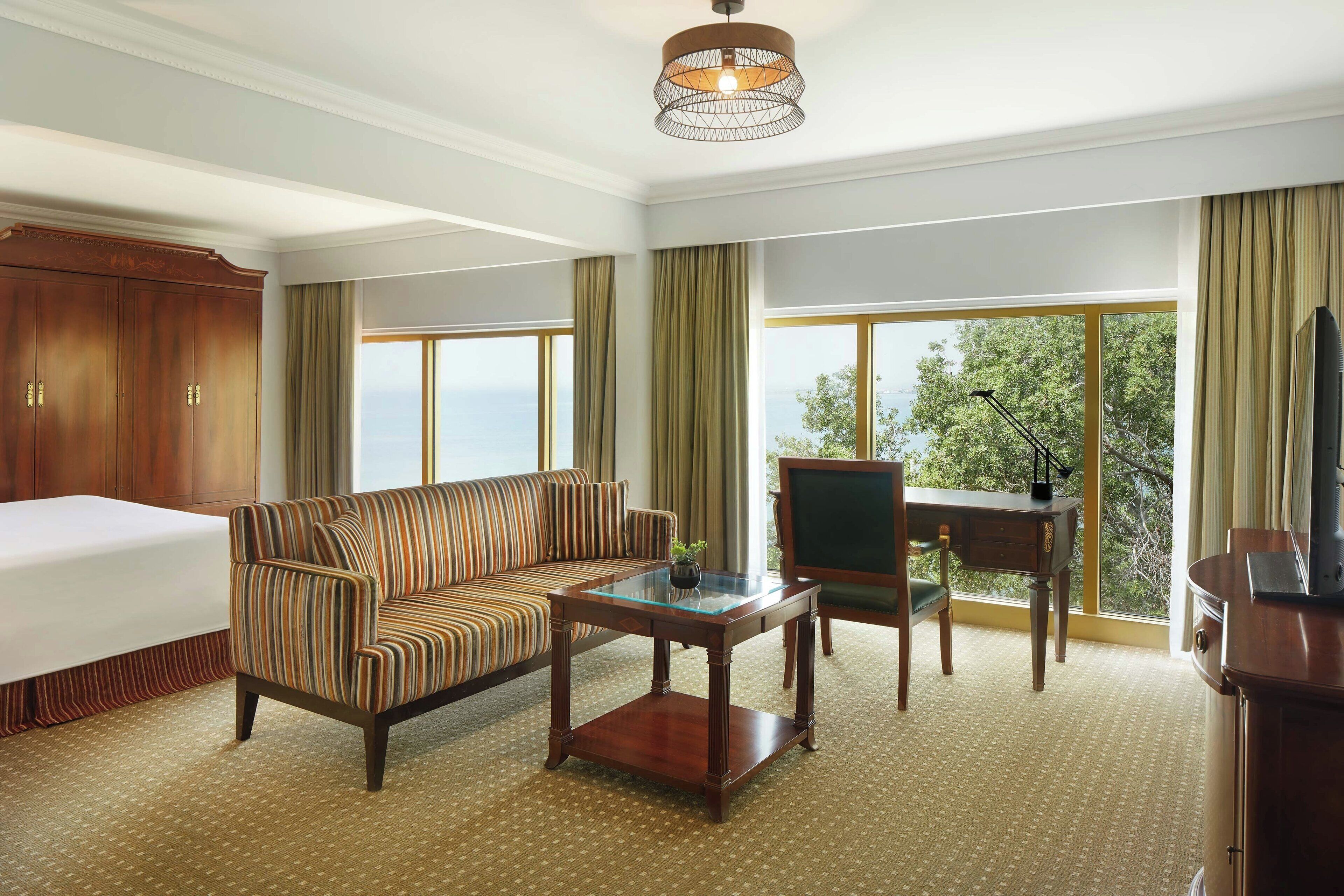 A hotel room with a bed, a striped sofa, a glass-top coffee table, a desk with a chair, and a television.