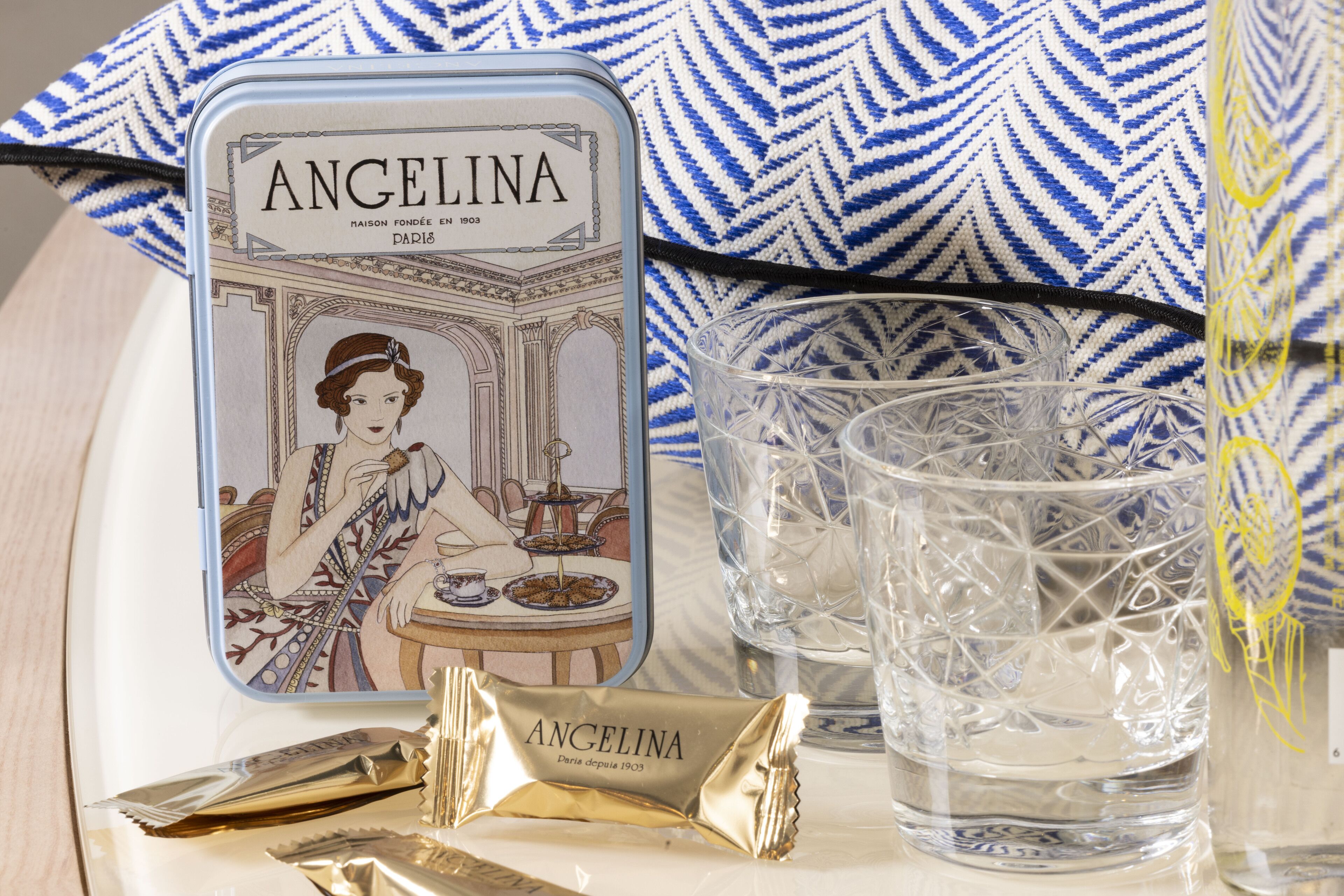 A tin of Angelina chocolates, two crystal glasses, and a patterned pillow.