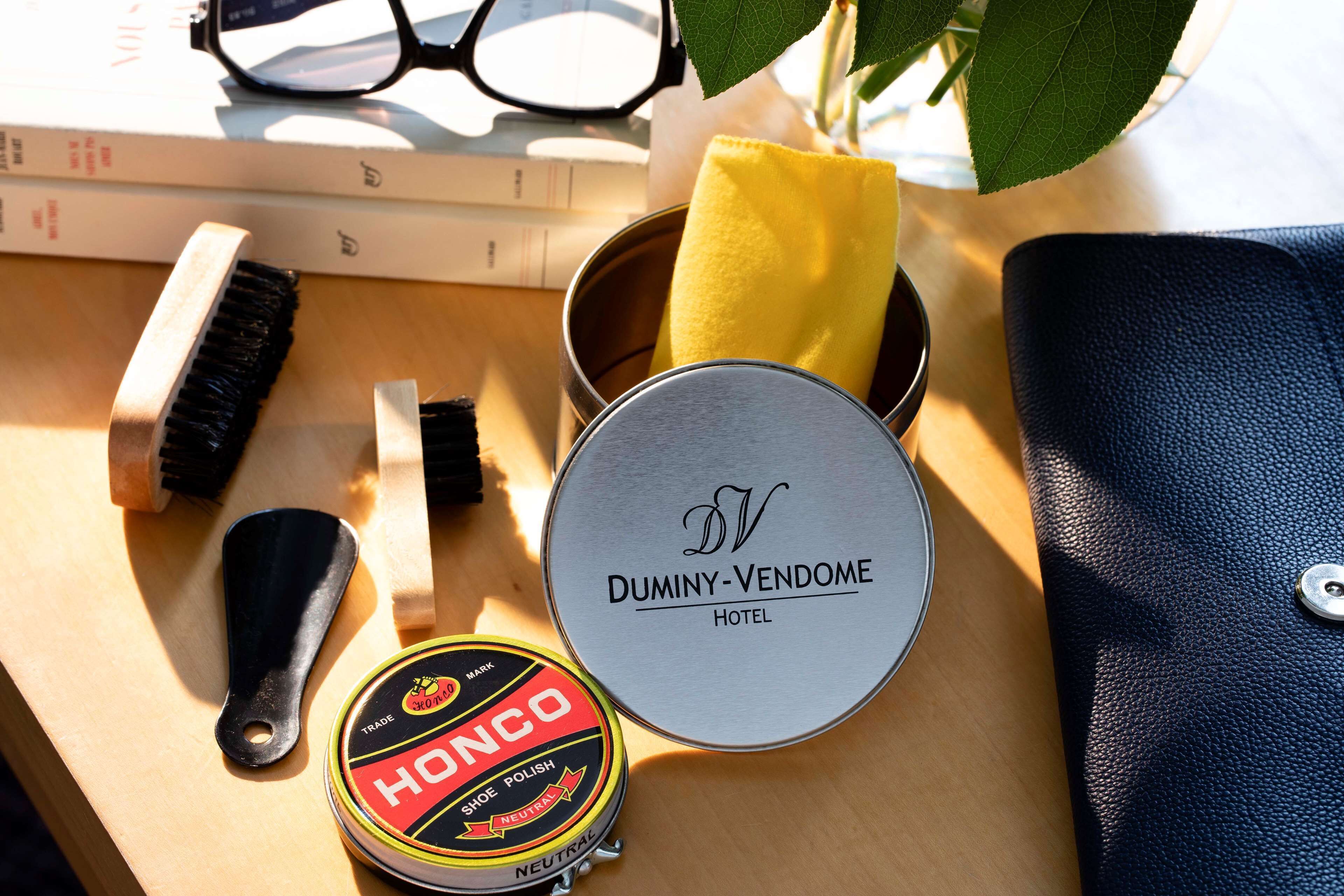Una superficie de madera con una lata redonda etiquetada como "DUMINY-VENDOME HOTEL", un pincel negro, un paño amarillo, un recipiente redondo con la marca "HONCO" y un par de gafas.