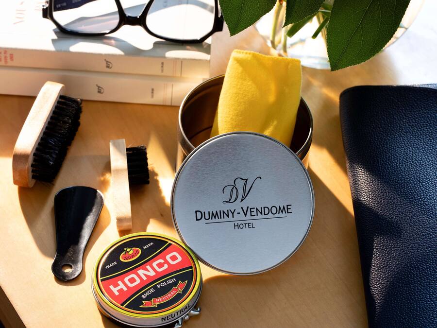 Una superficie de madera con una lata redonda etiquetada como "DUMINY-VENDOME HOTEL", un pincel negro, un paño amarillo, un recipiente redondo con la marca "HONCO" y un par de gafas.