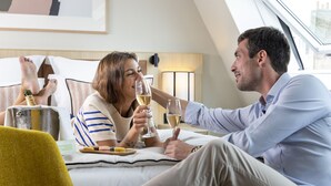 Una pareja sentada en una cama de hotel, sosteniendo copas de vino, con vistas a la ciudad a través de un tragaluz.