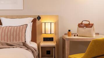 A hotel room with a bed, a bedside table with a lamp, a chair, and a handbag on the table.
