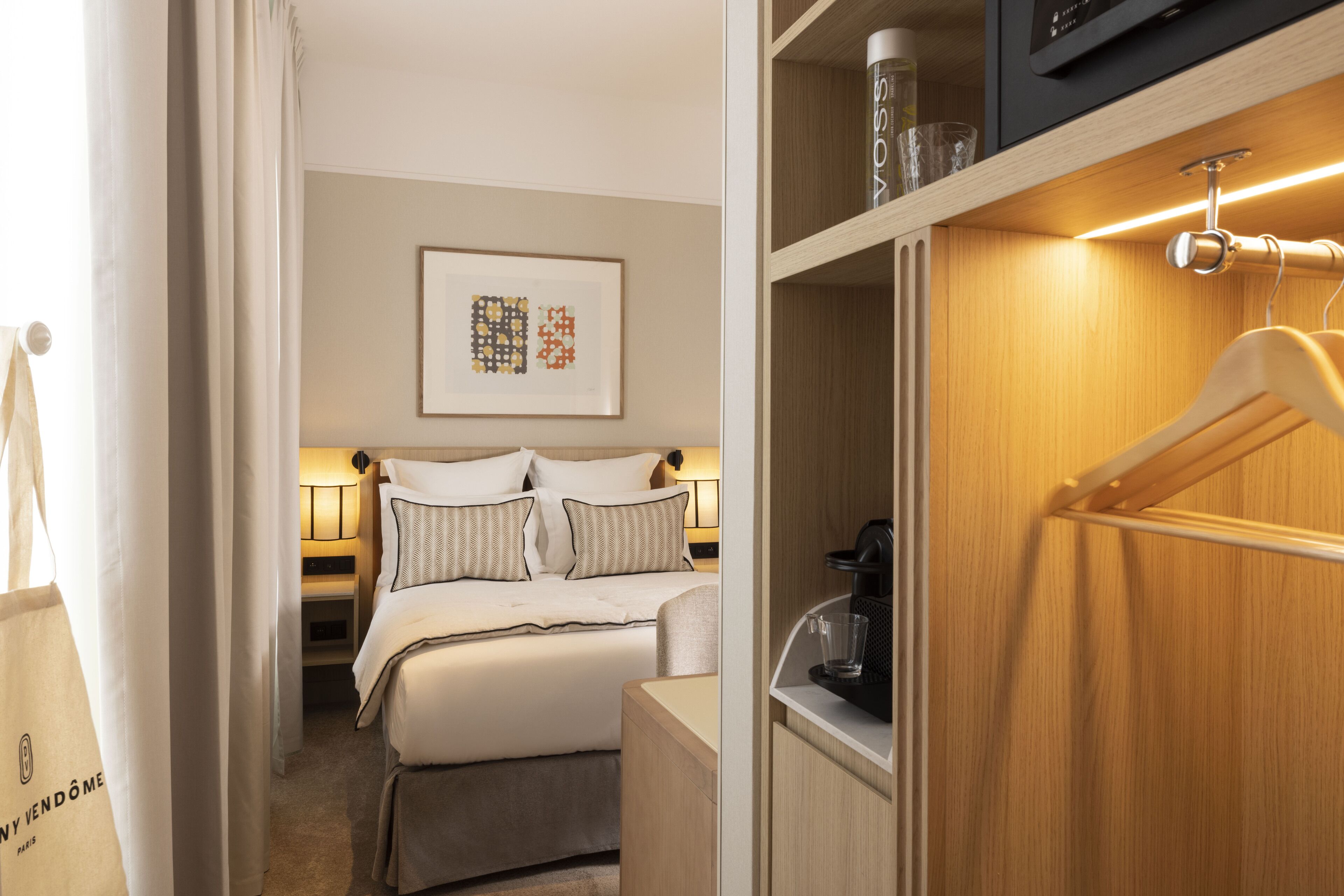 Quarto de hotel com uma cama bem arrumada, mesa de cabeceira com abajur, quadro na parede e guarda-roupa de madeira com cabides.