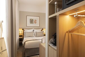 A hotel room with a neatly made bed, a bedside table with a lamp, a framed artwork on the wall, and a wooden wardrobe with a hanging rack.