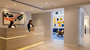 Un área de recepción de hotel moderna con un mostrador de madera, un cuadro abstracto grande y un interior luminoso con una alfombra azul estampada.