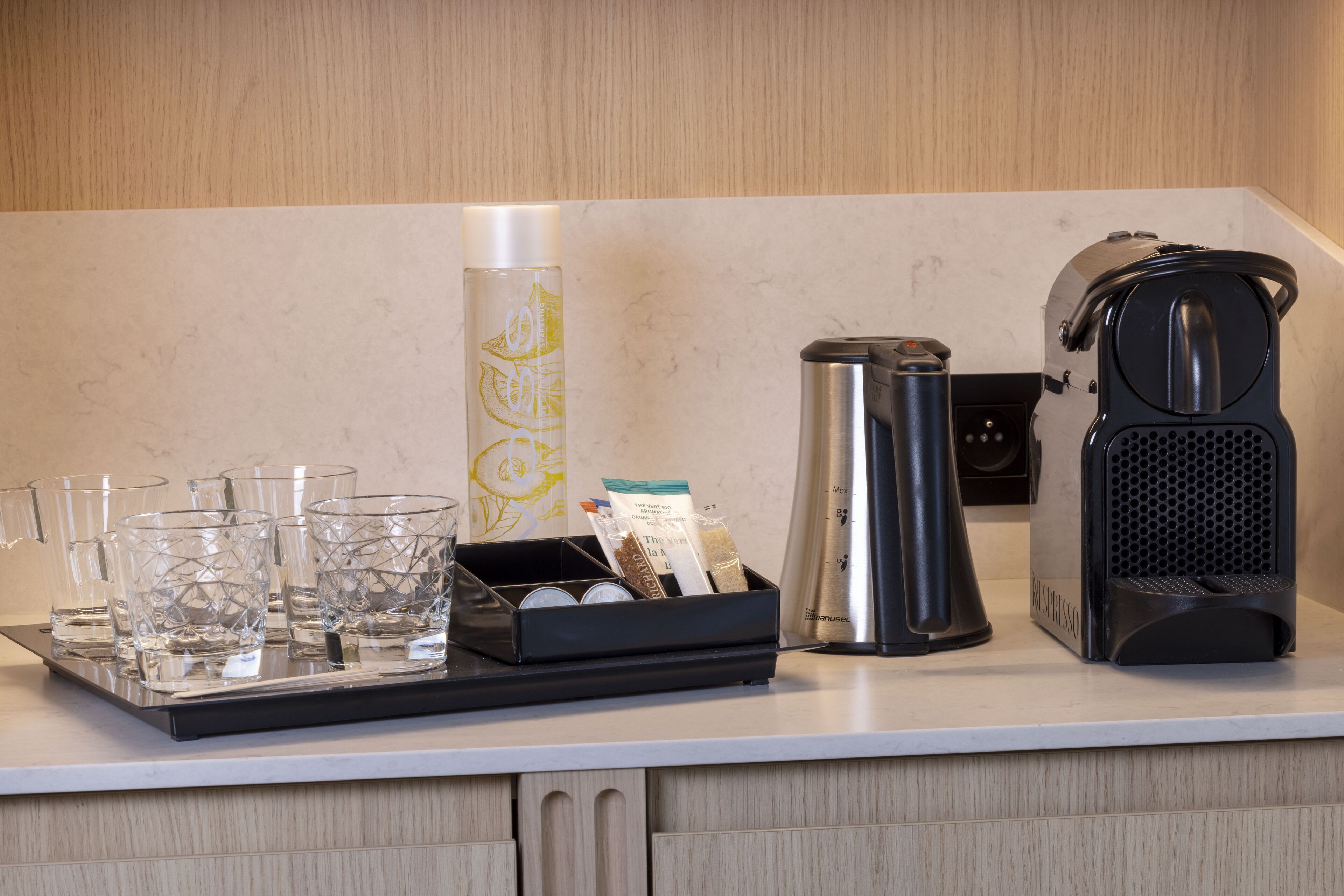 Een keukenblad met een koffiezetapparaat, een waterkoker, een dienblad met glazen en een fles, en een houder voor koffiecups.
