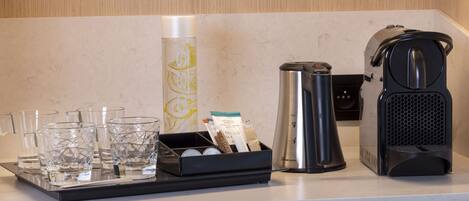 A kitchen counter with a coffee maker, a kettle, a tray with glasses and a bottle, and a coffee pod holder.