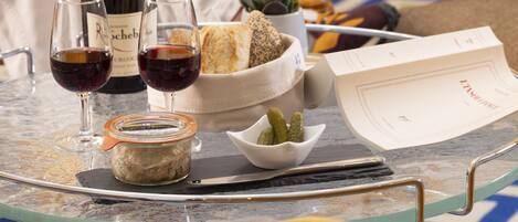 A table with a bottle of wine, two glasses of red wine, a jar of spread, a bowl of pickles, a bread basket, a small plant, and a menu on a glass table.
