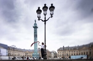 A person standing next to a tall, ornate street lamp with three lights, in front of a historic building with a statue on top.