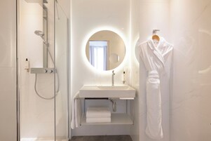 A modern bathroom with a glass shower enclosure, a white sink with a mirror above it, and a white robe hanging on a towel rack.