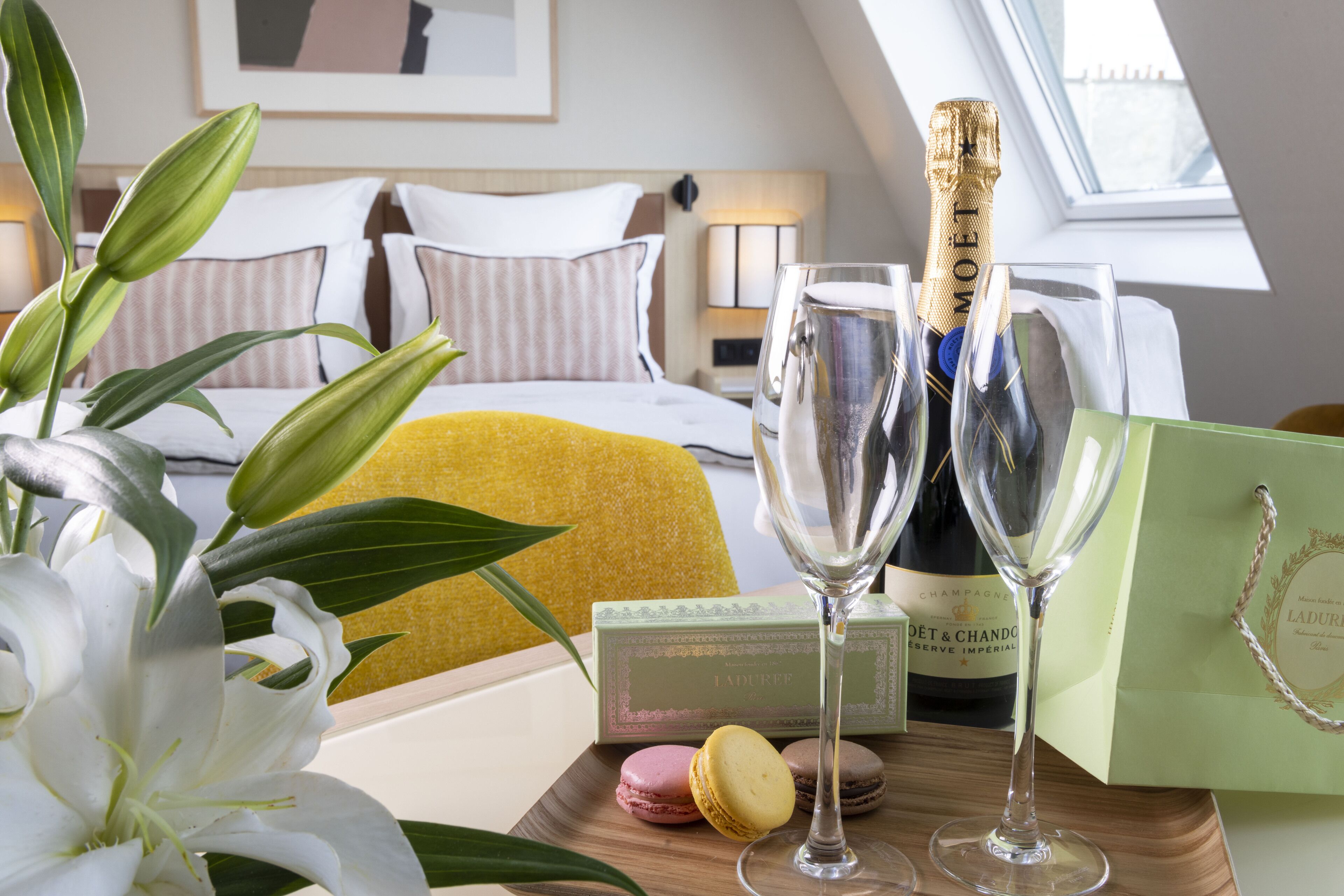 A hotel room with a bed, a nightstand, a window, and a table with a bottle of champagne, two glasses, a box of macarons, and a gift bag.