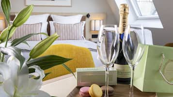 A hotel room with a bed, a nightstand, a window, and a table with a bottle of champagne, two glasses, a box of macarons, and a gift bag.