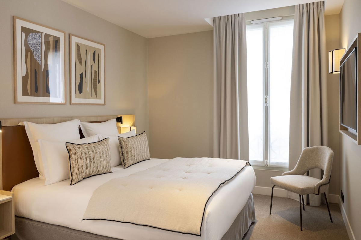 A hotel room with a large bed, a bedside table, a chair, a TV, and a window with curtains.