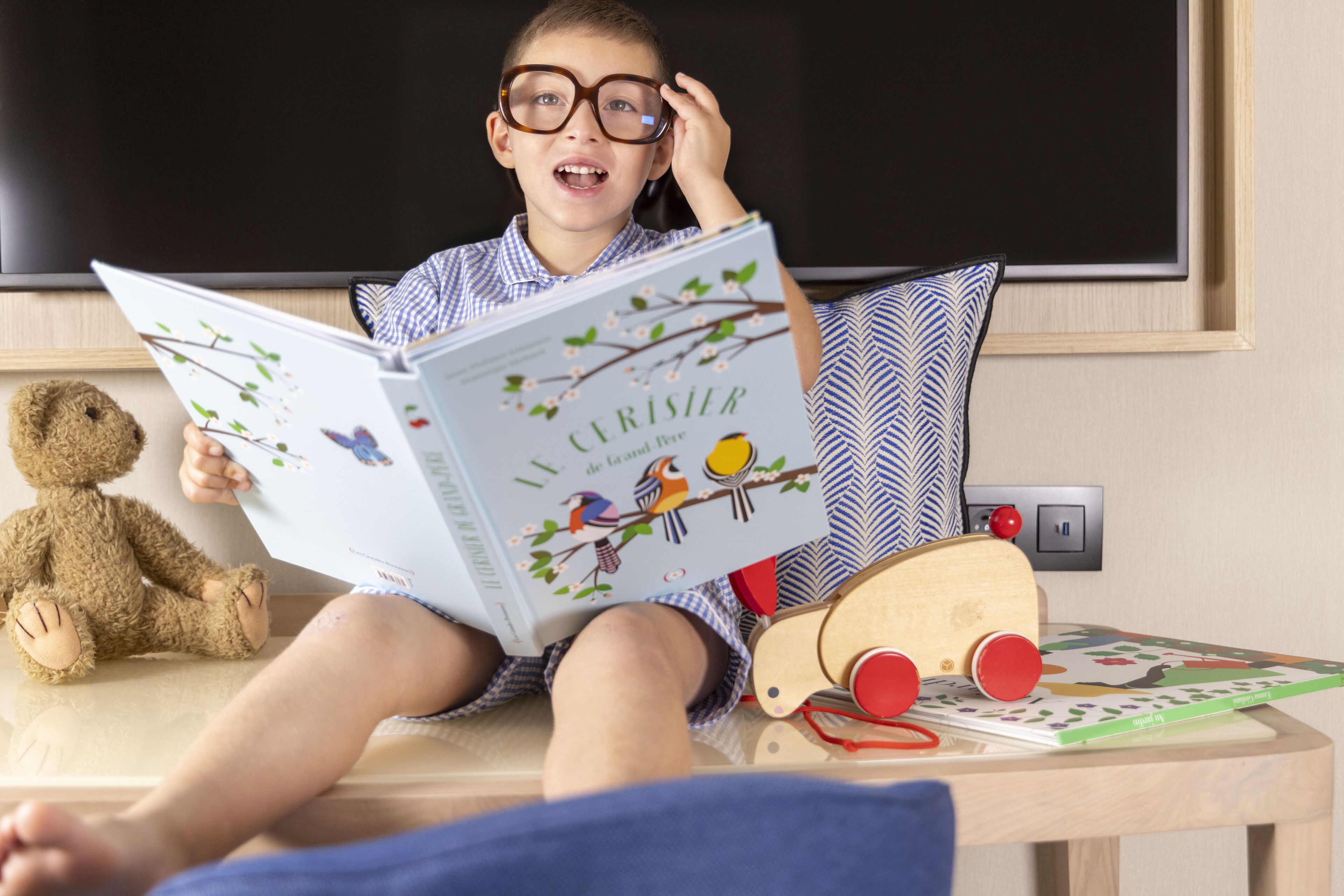 Un niño leyendo un libro en una habitación con una pizarra, un osito de peluche y un juguete de madera sobre una mesa.
