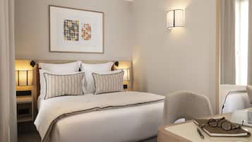 A hotel room with a neatly made bed, bedside lamps, a desk with a chair, and a framed artwork on the wall.