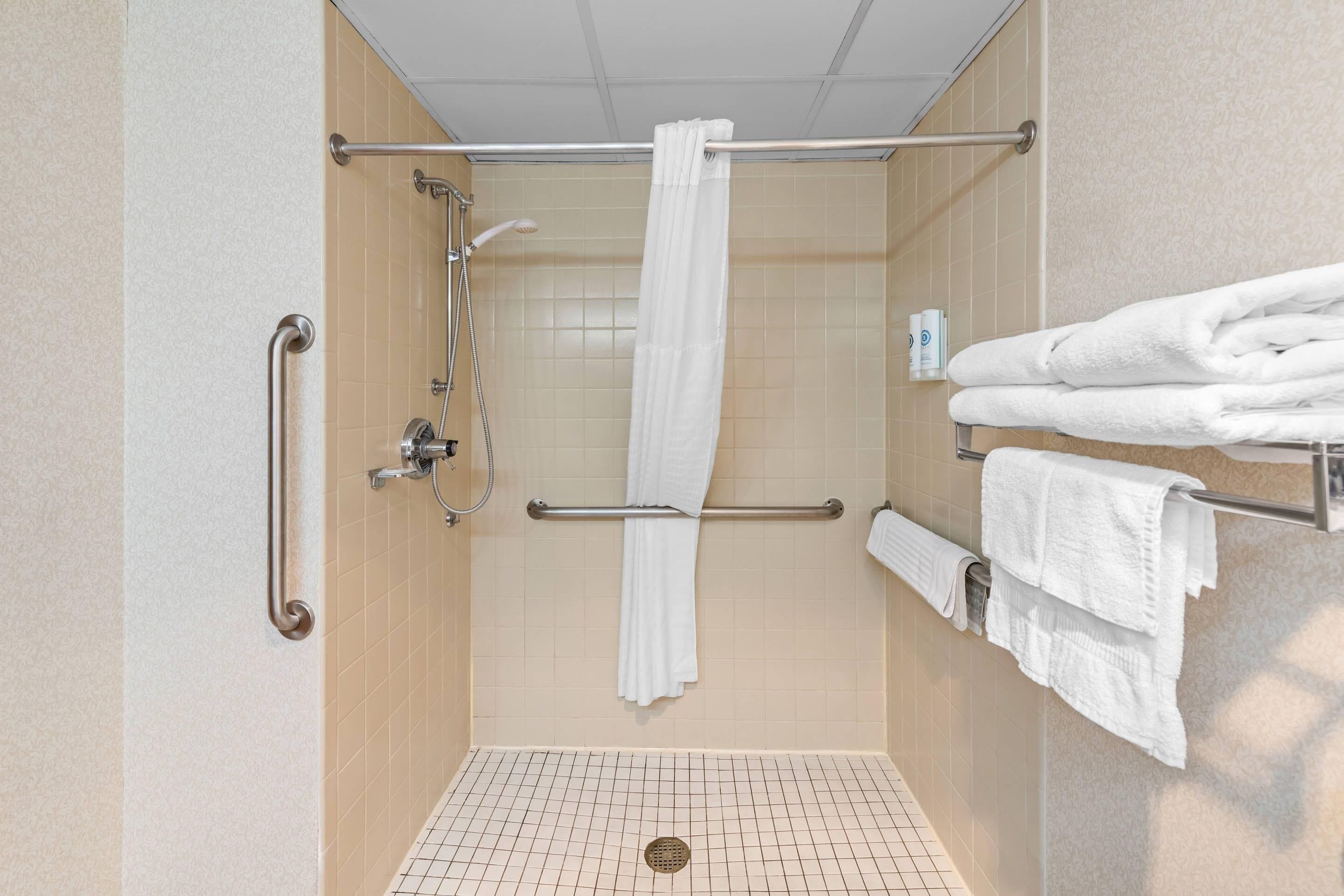 A shower area with a white shower curtain, grab bars, and a towel rack with folded towels.