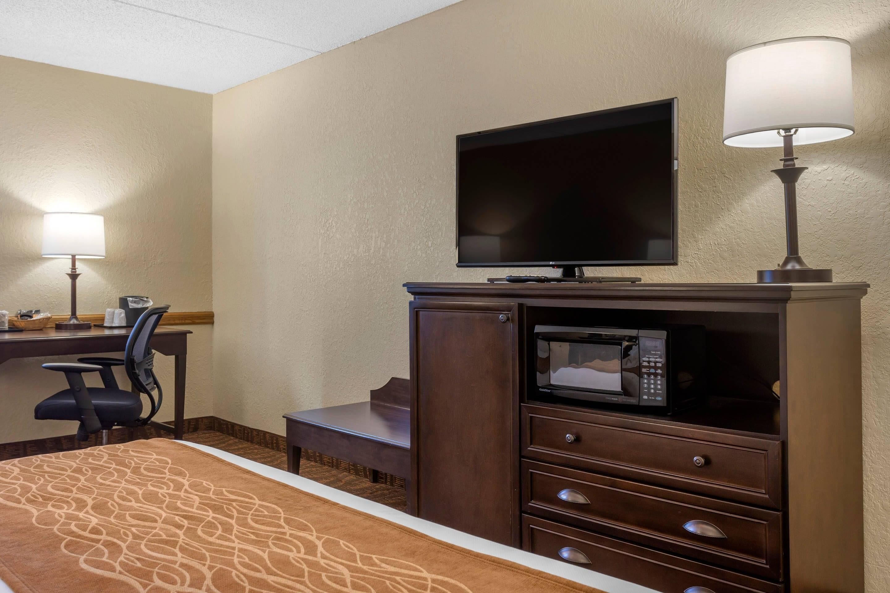 A hotel room with a bed, a desk with a chair, a television, a microwave, and two lamps.