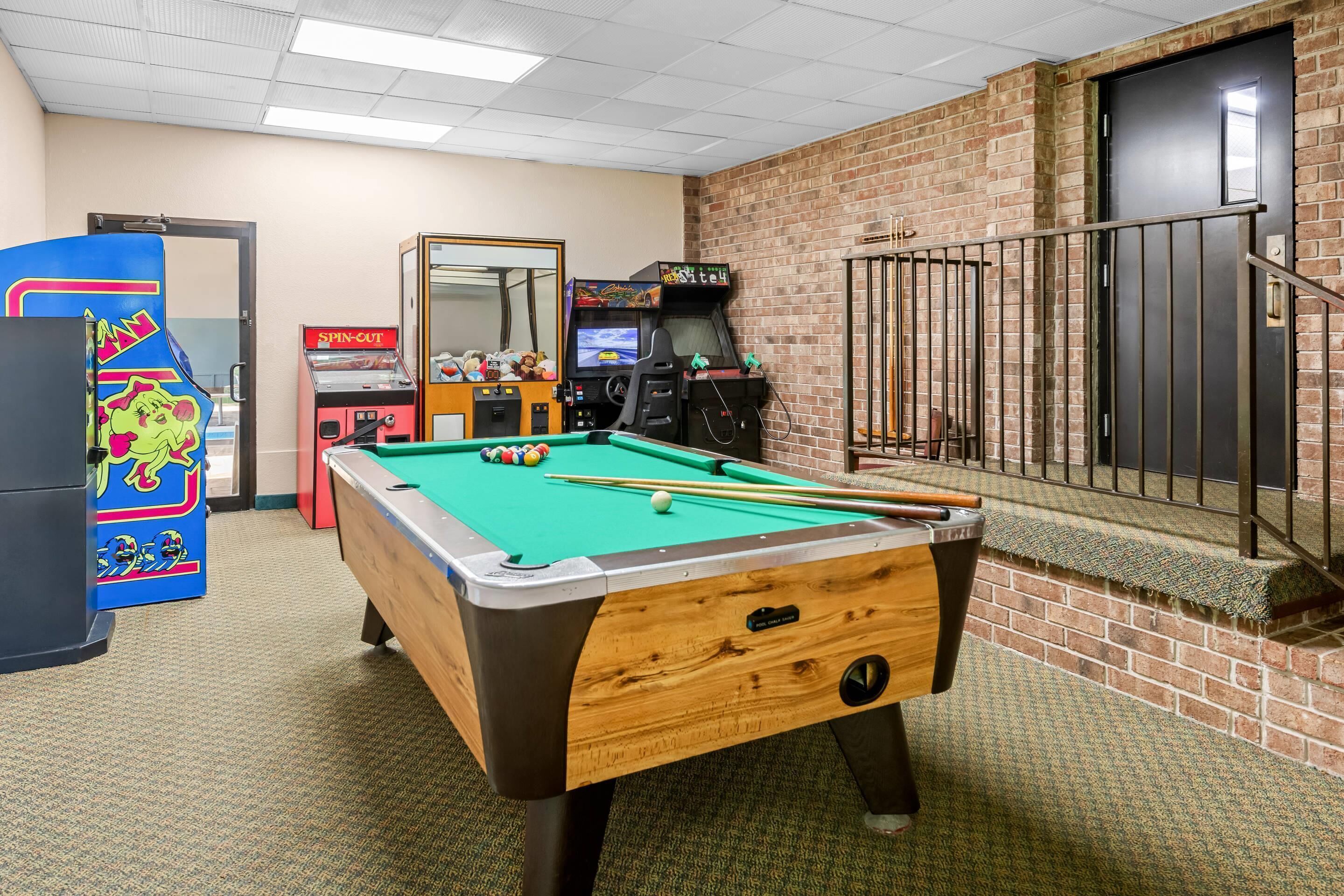 A pool table in a room with a brick wall, a door, and arcade games.