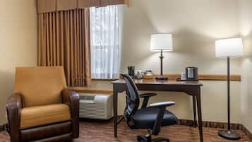 A hotel room with a brown leather chair, a desk with a lamp, a black office chair, and an air conditioner.