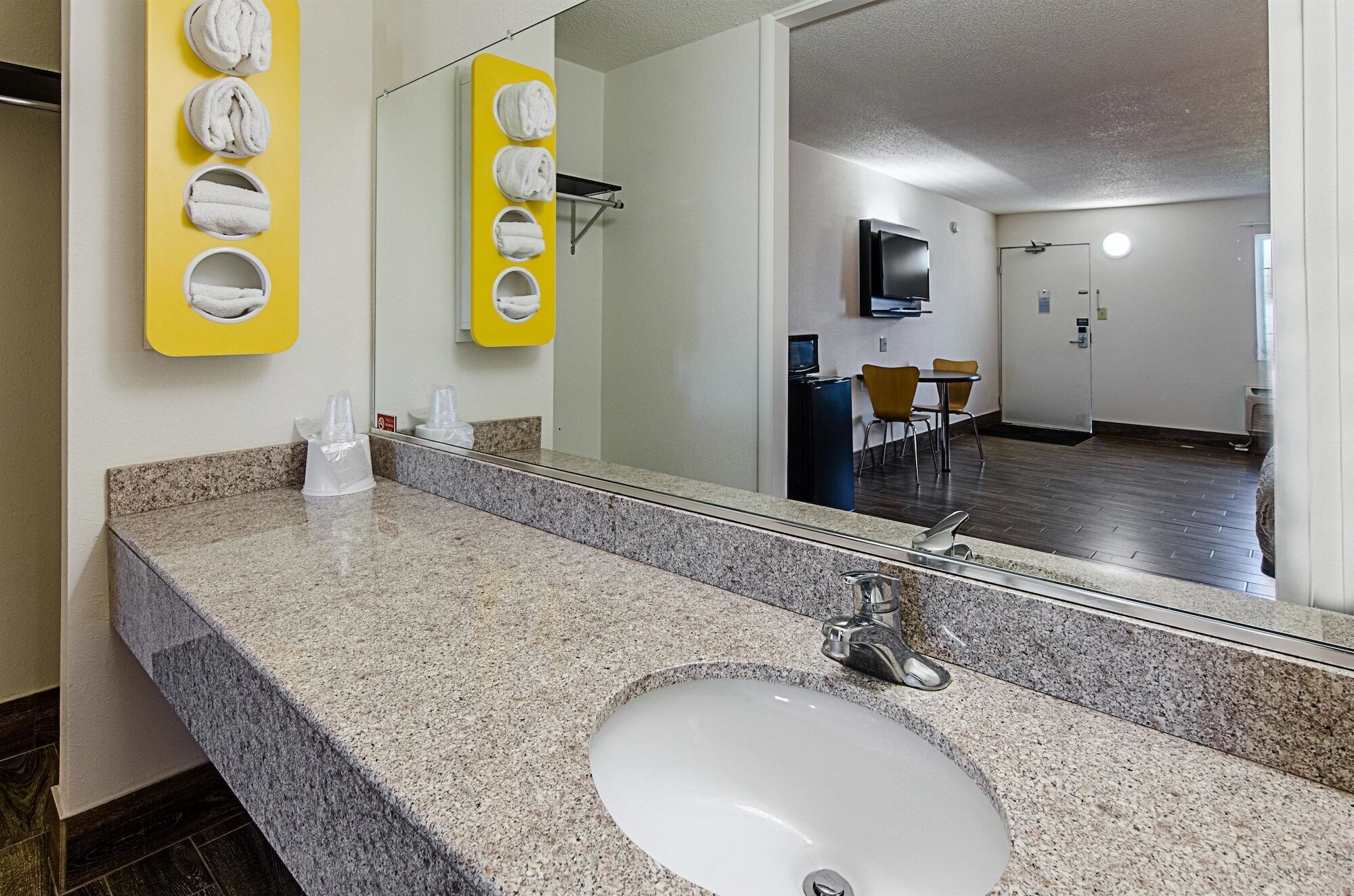 A hotel bathroom with a granite countertop, a sink, and a mirror. There are towels hanging on a yellow rack, and a chair is visible in the background.
