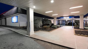 Entrada de un edificio moderno con cartel de recepción, un gran letrero iluminado con el número 6 y un área ajardinada bien cuidada.