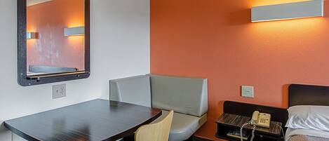 Habitación de hotel con escritorio, una silla, un banco, una cama, una mesita de noche con teléfono y una lámpara de pared.