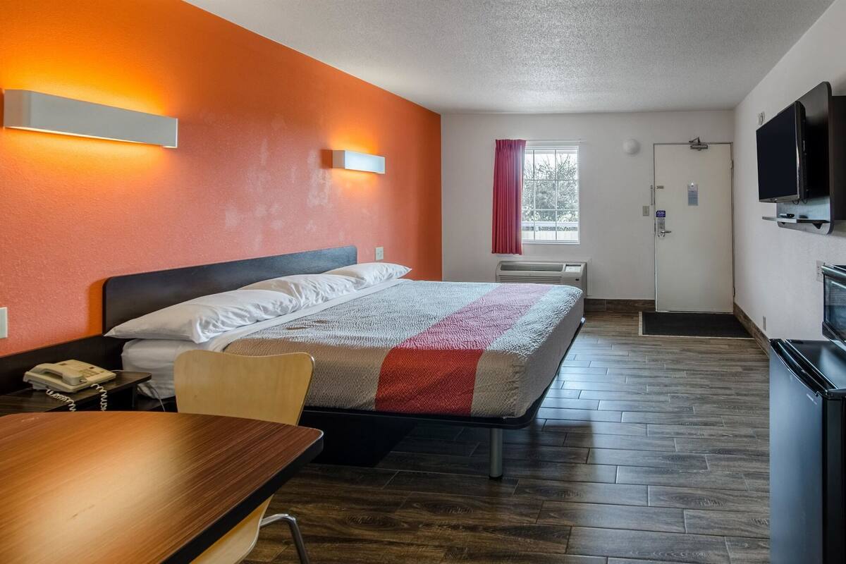 A hotel room with a bed, a desk, a chair, a TV, and a kitchenette.