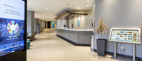 Un hall d’hôtel moderne doté d’un kiosque numérique, d’un comptoir d’accueil et d’un escalier menant à un autre étage.