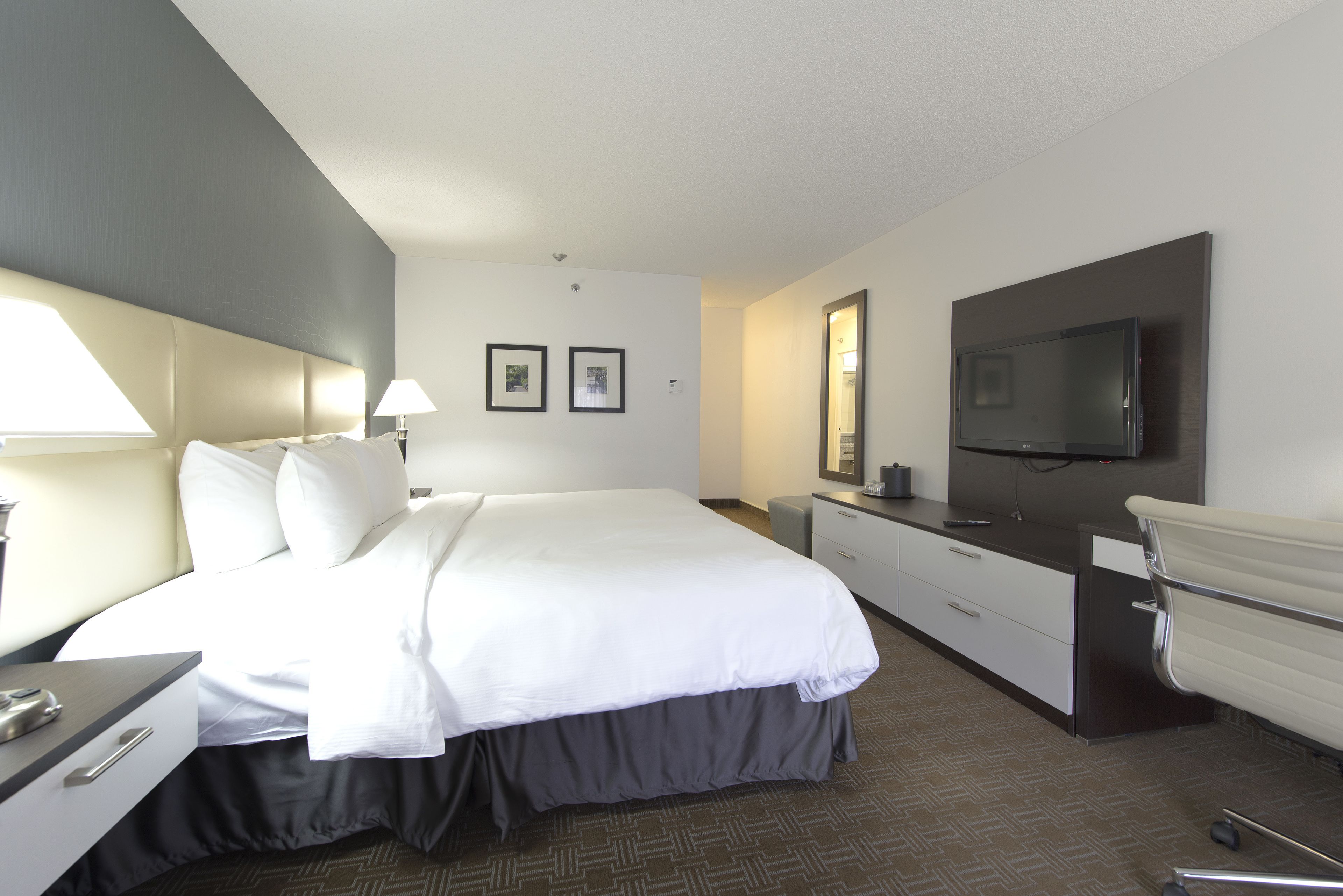 Una habitación de hotel con una cama grande, un escritorio con lámpara, un aparador, un espejo y un televisor instalado en la pared.