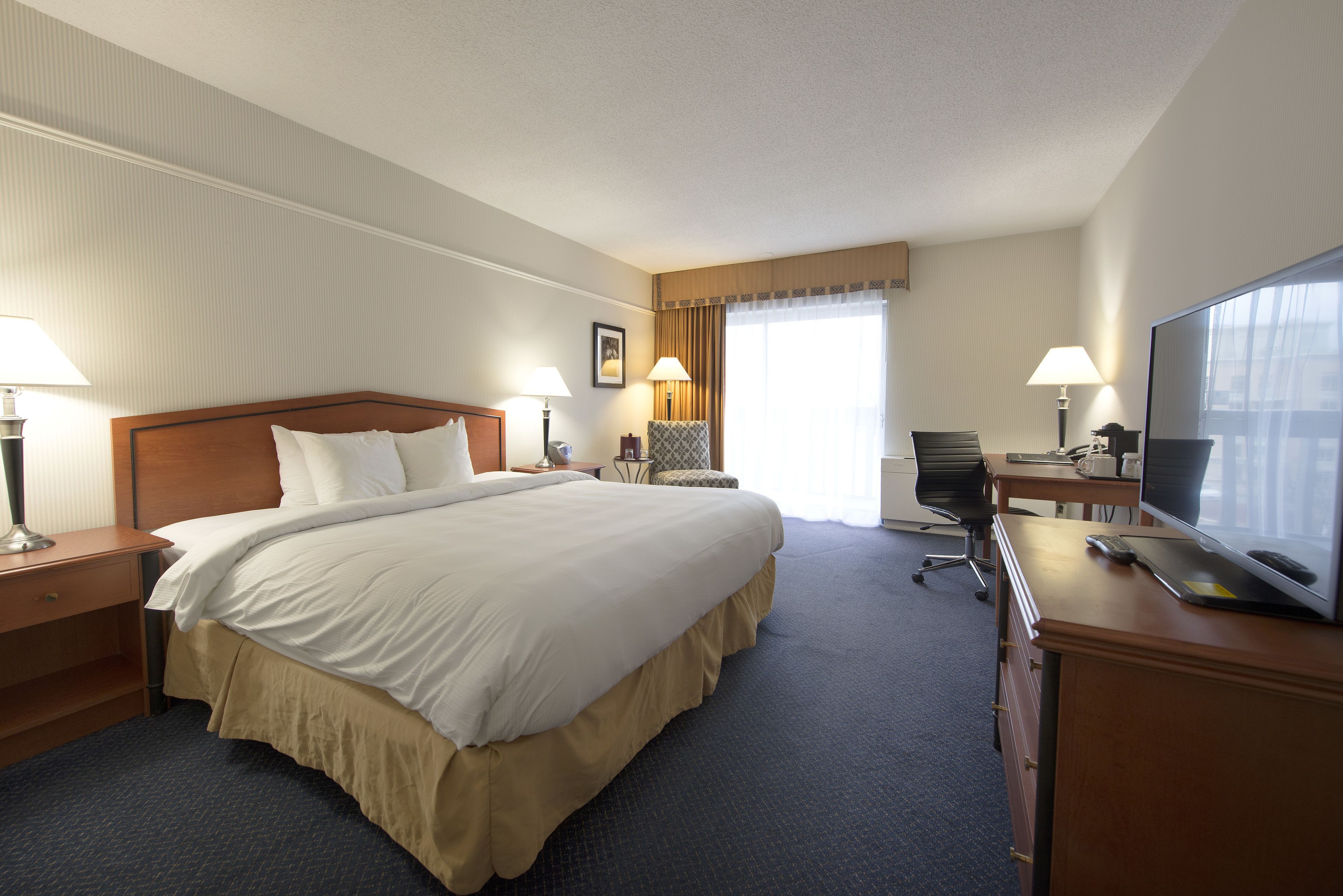 Una habitación de hotel con una cama grande, dos mesitas de noche con lámparas, un escritorio con una computadora y un televisor, y una silla.