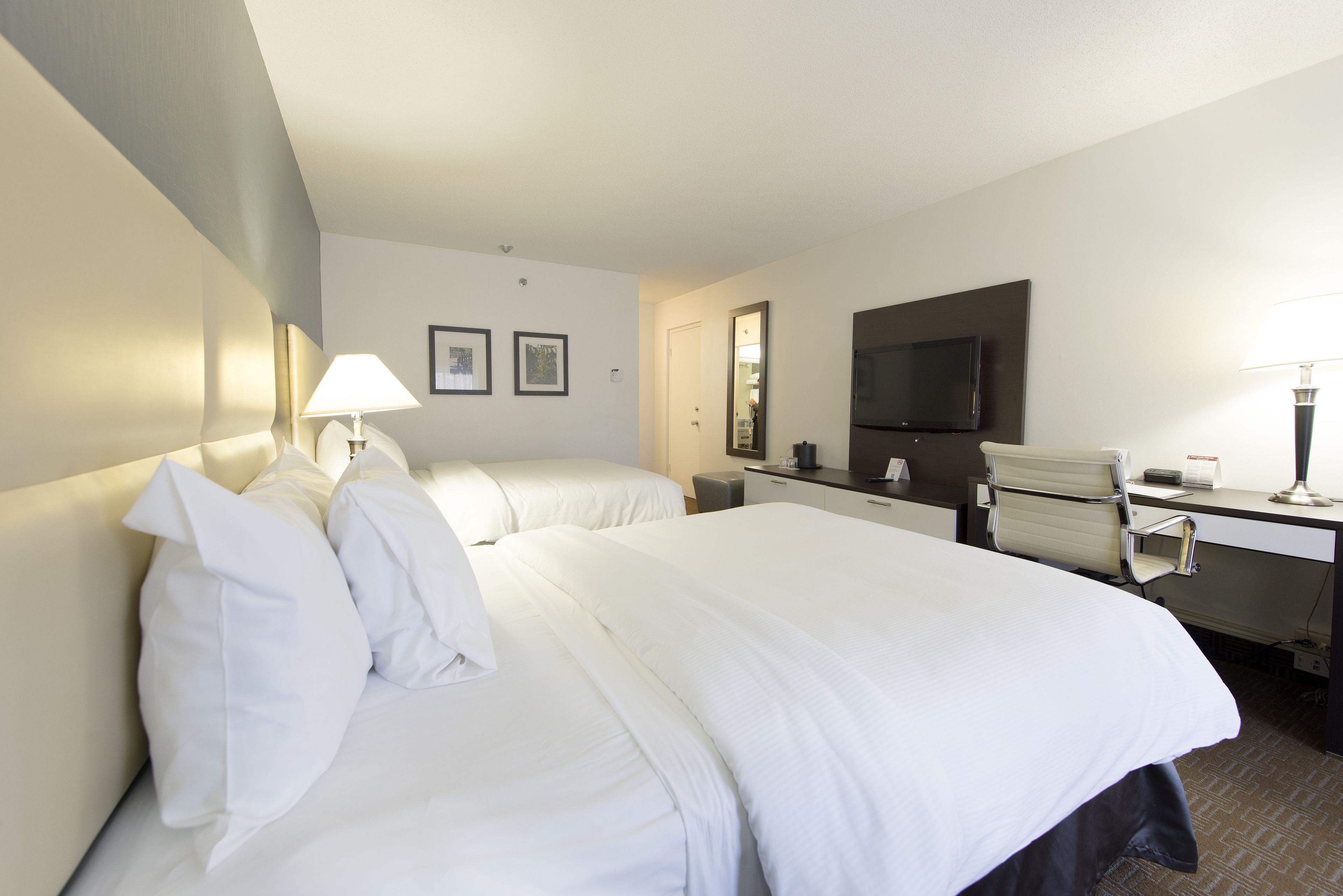 A hotel room with two beds, a desk, a chair, a TV, and a lamp.