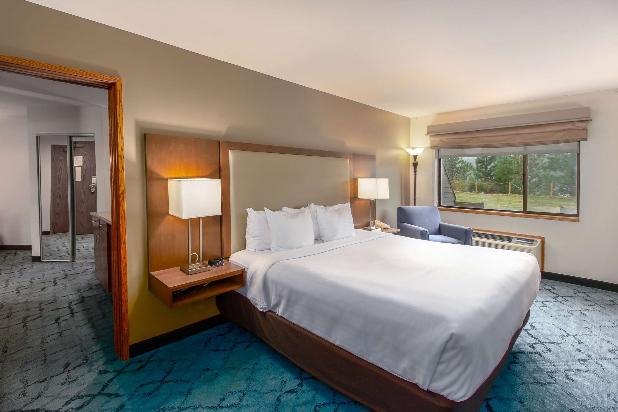A hotel room with a large bed, two bedside lamps, a desk with a chair, a window with a view of the outdoors, and a mirror on the left wall.