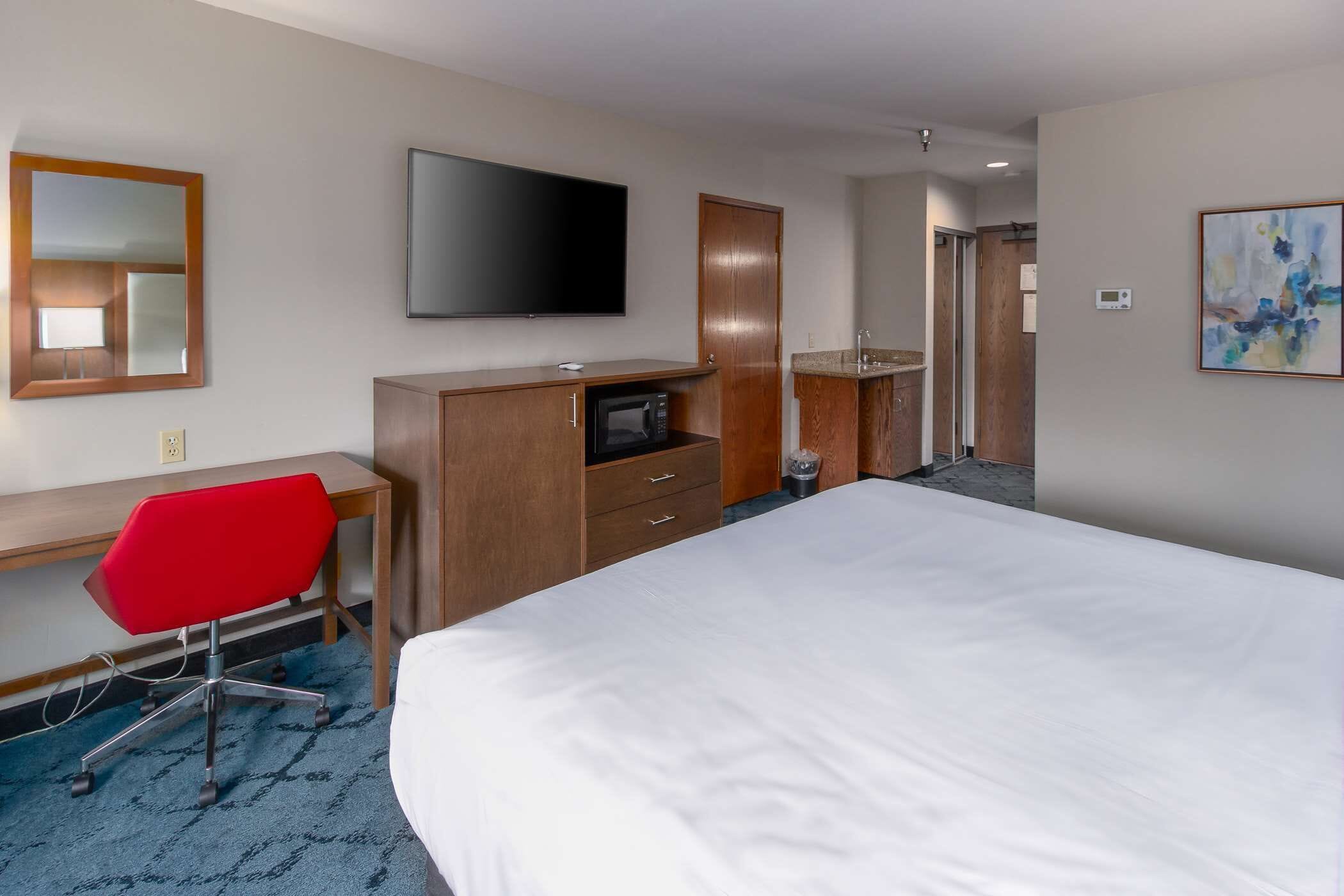 A hotel room with a bed, a desk with a red chair, a television, a microwave, and a bathroom visible in the background.
