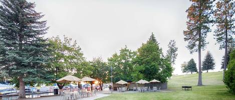 An outdoor patio area with tables and chairs, surrounded by trees and a grassy hill.