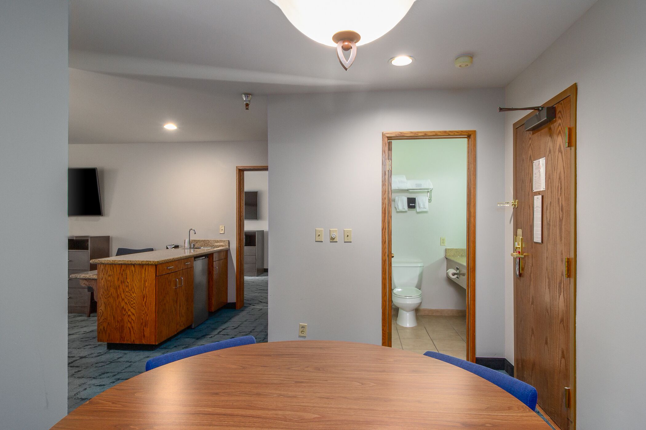 A hotel room with a wooden dining table, a kitchenette, a bathroom with a toilet and sink, and a mirror.