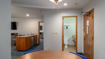 A hotel room with a wooden dining table, a kitchenette, a bathroom with a toilet and sink, and a mirror.