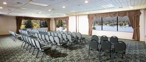 Un salón de conferencias con filas de sillas, ventanas amplias con cortinas y vista a un lago.
