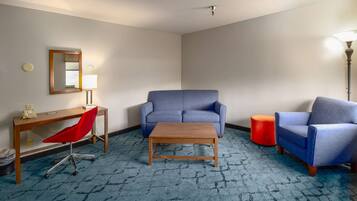 A hotel room with a blue carpet, a wooden desk with a red chair, a blue sofa, a wooden coffee table, and a red ottoman.