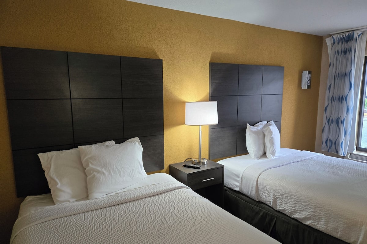 Une chambre d’hôtel avec deux lits, une table de chevet, une lampe et une fenêtre avec des rideaux.