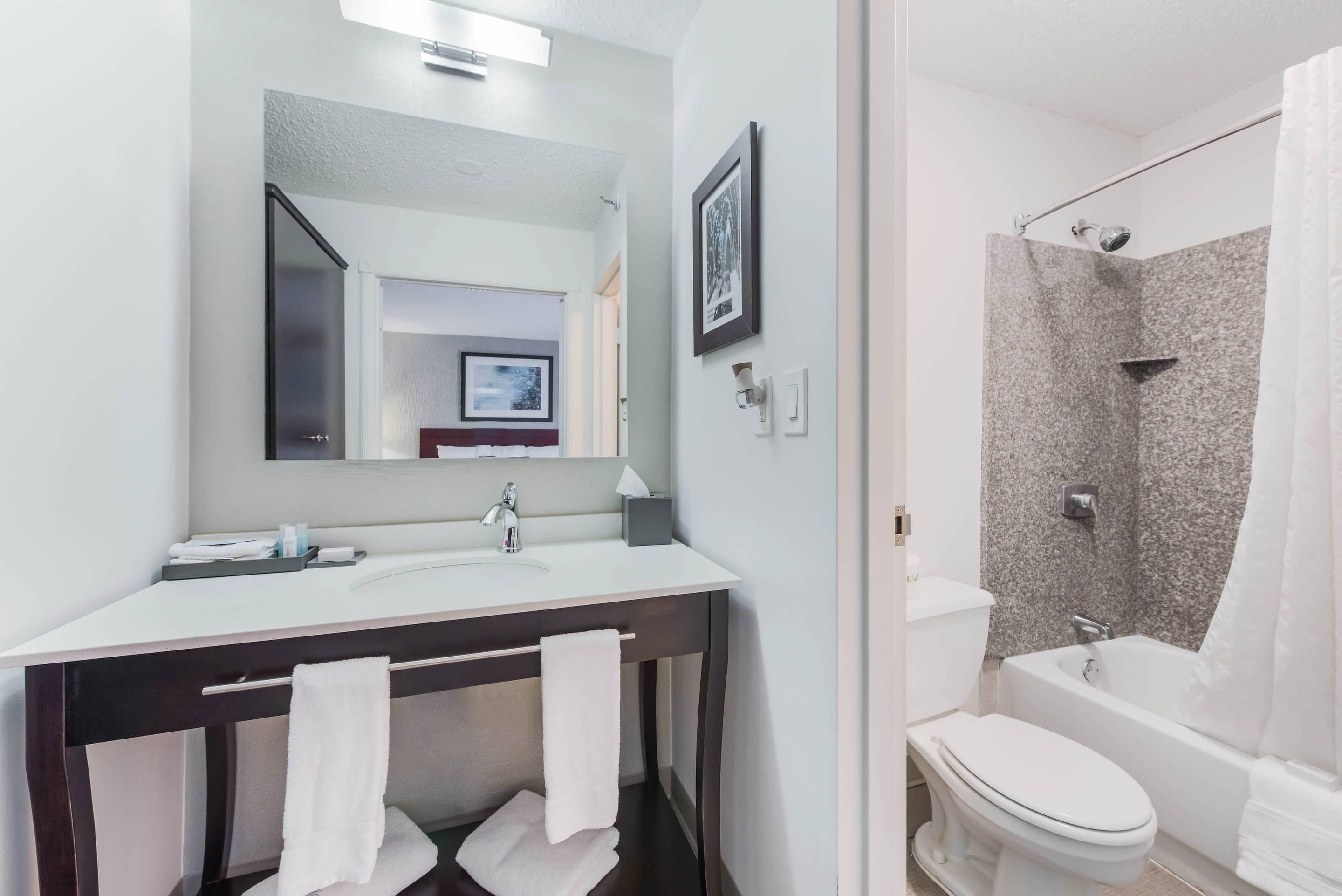 A modern bathroom with a white sink, dark wooden vanity, white towels, a large mirror, a framed picture, and a corner shower with a white bathtub.